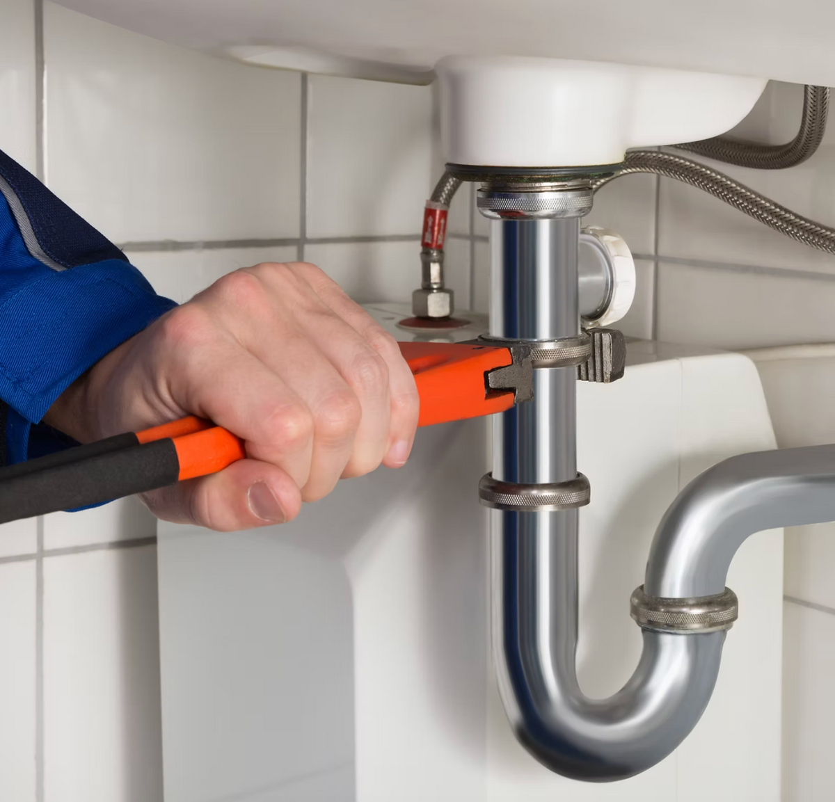 A close-up of a plumber's hand using orange-handled pipe wrenches to tighten a metal pipe clamp under a sink, with white tiled walls in the background.